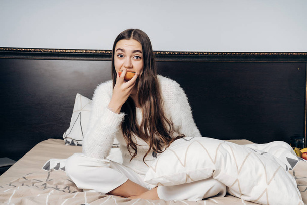 encantadora chica de pelo largo se sienta en la cama, desayunos una deliciosa manzana útil
 - Foto, Imagen