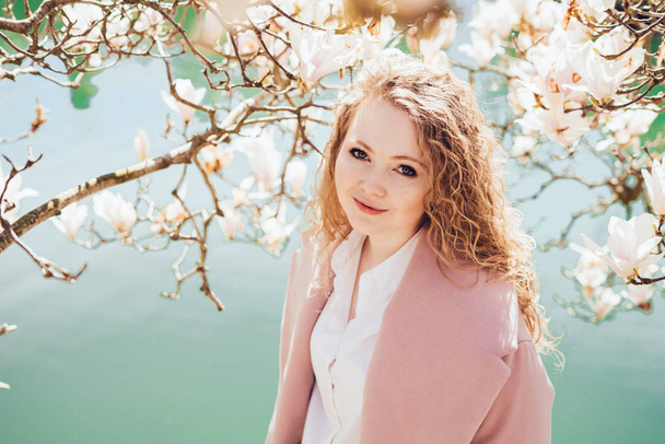 elegant young girl in a pink coat enjoys blooming fragrant magnolia in the park, by the pond - Foto, afbeelding
