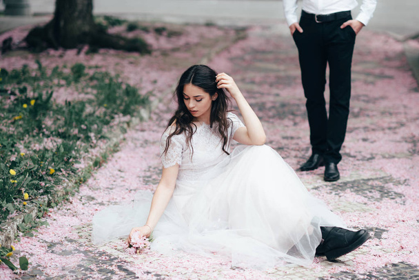 Portrait of a young beautiful brunette girl in white wedding dress sitting on a grass in a blooming spring garden. - Fotó, kép