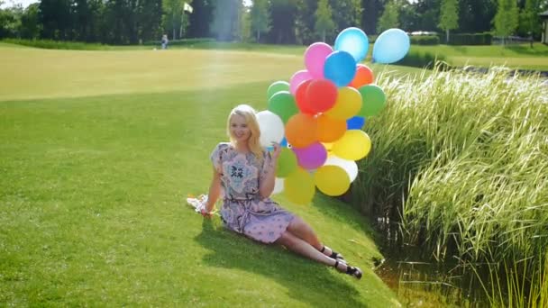 Zeitlupenaufnahmen einer glücklichen jungen Frau im Kleid, die mit Luftballons auf Gras sitzt - Filmmaterial, Video