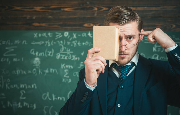 Closeup portrait of student holding book in front of his face and pointing at his head with other hand. Thoughtful intelligent man in suit standing in classroom - 写真・画像