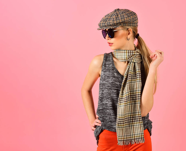 Side view skinny blond model in tartan beige cap and scarf and black sunglasses isolated on pink background. Girl in bright orange pants playing with her ponytail - Foto, afbeelding