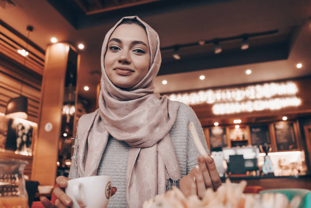 a cute Muslim girl wearing a hijab sits in an oriental cozy cafe, dines and looks at the camera - Fotoğraf, Görsel