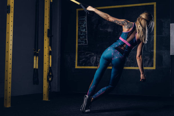 Mulher loira musculosa bonita em sportswear com uma tatuagem em seu braço fazendo exercício com sistema TRX no ginásio
. - Foto, Imagem