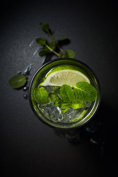 Glass of iced Mojito cocktail and ingredients,  lime, ice and mint leaves on black background, cold summer drink  - Фото, зображення