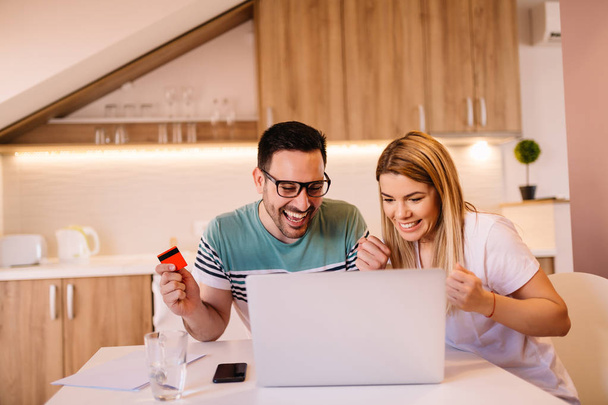 Onnellinen pari tekee verkko-ostoksia luottokortilla ja kannettavalla tietokoneella istuessaan modernissa keittiössä
 - Valokuva, kuva