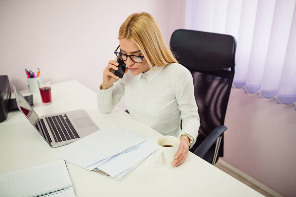 Blond podnikatelka s telefonní hovor při sezení v kanceláři před laptop a poznámky - Fotografie, Obrázek