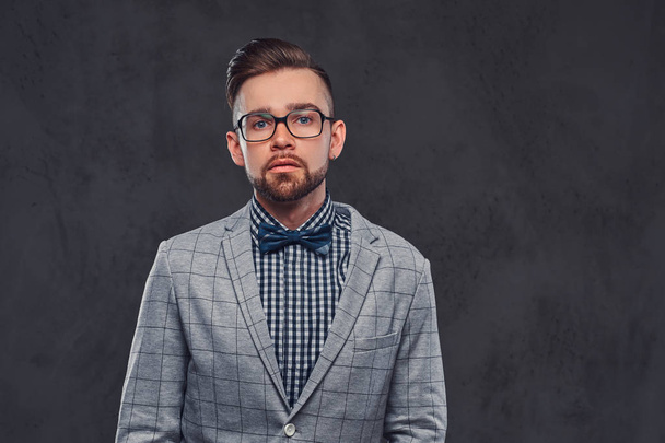Homem barbudo elegante confiante com penteado e óculos em um terno cinza retro elegante e gravata borboleta, posando em um estúdio
. - Foto, Imagem