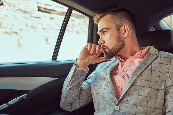Joven empresario con estilo de éxito en un traje gris y camisa rosa, montado en un asiento trasero en un coche de lujo
. - Foto, Imagen