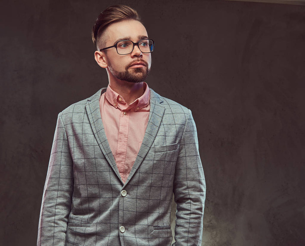 Retrato de un hombre barbudo con estilo y confianza con peinado y gafas en un traje gris y camisa rosa, posando en un estudio
. - Foto, imagen