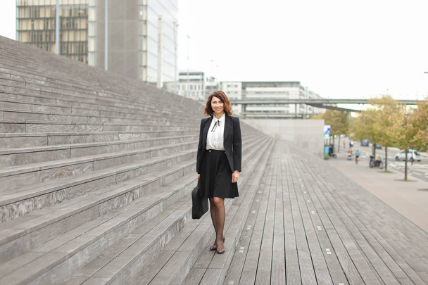 American businesswoman standing on stairs with bag and high buildings in background. - 写真・画像