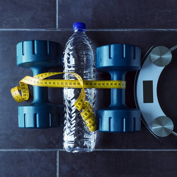 Dumbbells lie next to a bottle of pure mineral water and electronic scales. Yellow measuring tape unites them. - Photo, Image