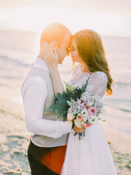 Cute portrait of the happy couple. The bride is petting the face of the groom at the background of the sunset in the sea. - Zdjęcie, obraz