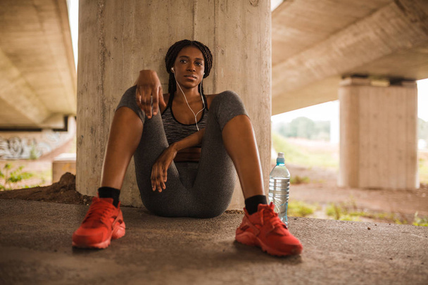 Young tired african american woman with earphones sitting on floor after running  - Fotografie, Obrázek