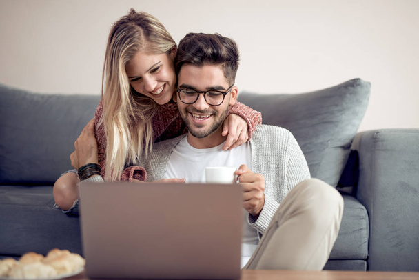 Όμορφο νεαρό ζευγάρι χρησιμοποιώντας ένα φορητό υπολογιστή, χαμογελώντας ενώ κάθεστε στον καναπέ στο σπίτι. Αγόρι εκμετάλλευση από ένα φλιτζάνι. - Φωτογραφία, εικόνα