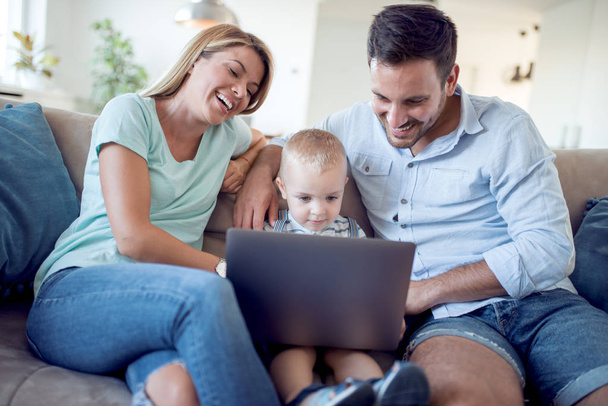 glücklicher junger Vater und Mutter, die den Laptop in der Hand halten und surfen, während sie mit ihrem Baby sitzen. - Foto, Bild