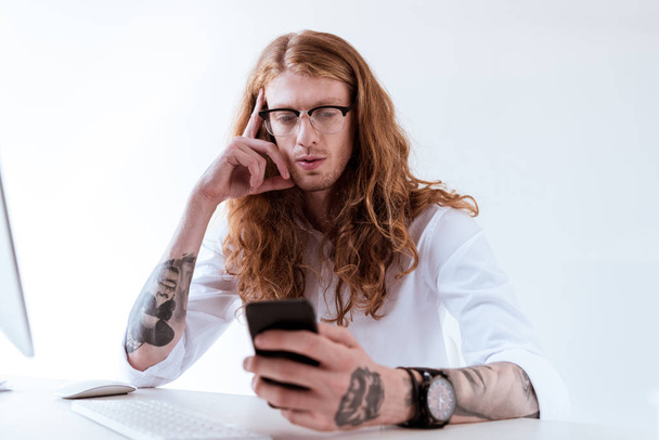 hombre de negocios tatuado con estilo con el pelo rizado usando el teléfono inteligente en la mesa en la oficina
 - Foto, Imagen
