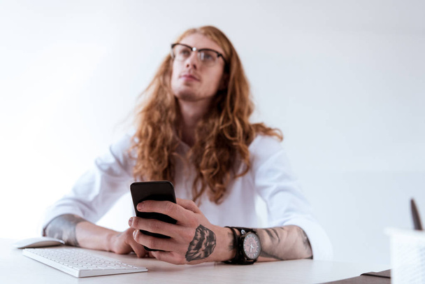 zamyšlený stylový Tetovaný podnikatel s kudrnatými vlasy drží smartphone a vyhledávání - Fotografie, Obrázek