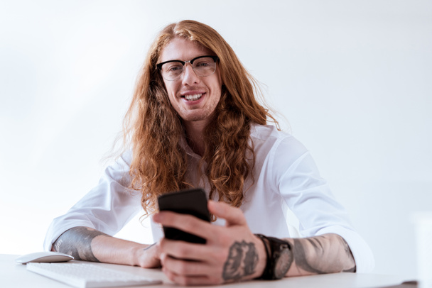 sonriente elegante hombre de negocios tatuado con pelo rizado celebración de teléfono inteligente
 - Foto, Imagen