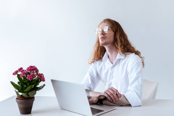 stijlvolle getatoeëerd zakenman met krullend haar aan tafel zitten met laptop en opzoeken - Foto, afbeelding