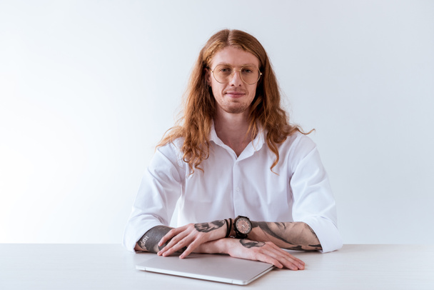 hombre de negocios tatuado con estilo con el pelo rizado sentado en la mesa con el ordenador portátil y mirando a la cámara aislada en blanco
 - Foto, Imagen