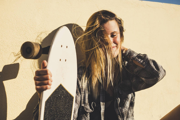 summer lifestyle beautiful blonde woman young sit down with her skateboard. smiling and touching her hair moved by the wind. freedom concept. - 写真・画像