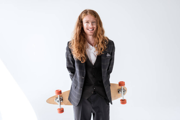 smiling stylish businessman with curly hair holding longboard above back - Foto, afbeelding