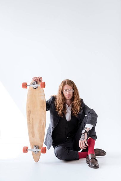 stylish businessman with curly hair sitting on floor and holding skateboard - Fotó, kép