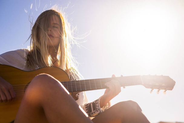 summer alternative vacation concept for beautiful blonde model female playing the guitar under a sunny day. sun backlight and girl immersed on her music. artist singer lifestyle - Photo, image