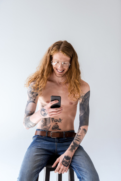gömleksiz gülümsüyor kıvırcık saçlı beyaz izole smartphone bakarak adam dövme - Fotoğraf, Görsel