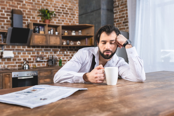 handsome loner businessman sitting with cup of coffee and sleeping at table at kitchen - Foto, Imagem