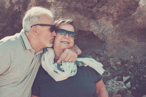 Счастливая пожилая пара в отпуске на Тенерифе в белых волосах и солнцезащитных очках улыбается и целует друг друга в щеку
 - Фото, изображение