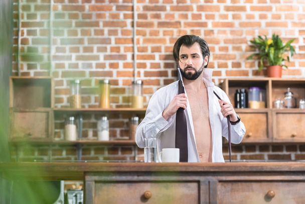 handsome loner businessman with unbuttoned shirt looking away at kitchen - Foto, afbeelding