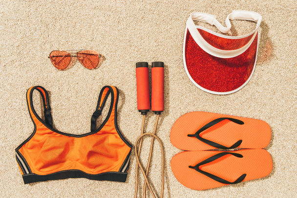 вид сверху на скакалку, спортивную одежду, солнцезащитные очки, колпачки и шлепанцы на песке
 - Фото, изображение