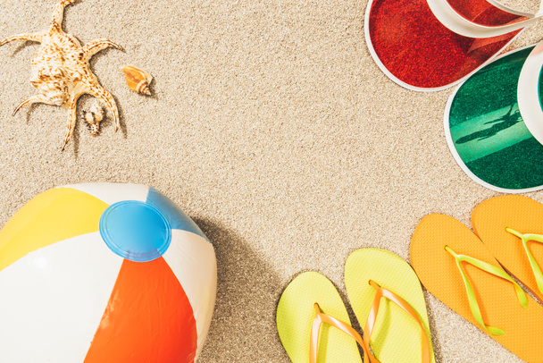 flat lay with colorful flip flops, beach ball, seashells and caps arranged on sand - 写真・画像