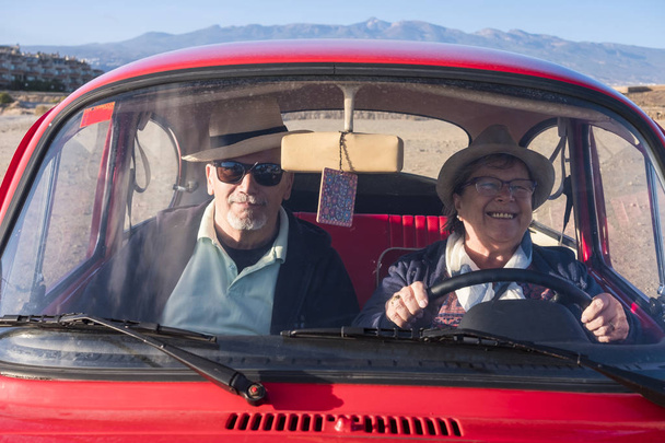 Régi piros autóval egy két idősebb férfi és nő, mosolyog, és ő néz rád. Mind a hat. Alternatív utazási koncepció. - Fotó, kép