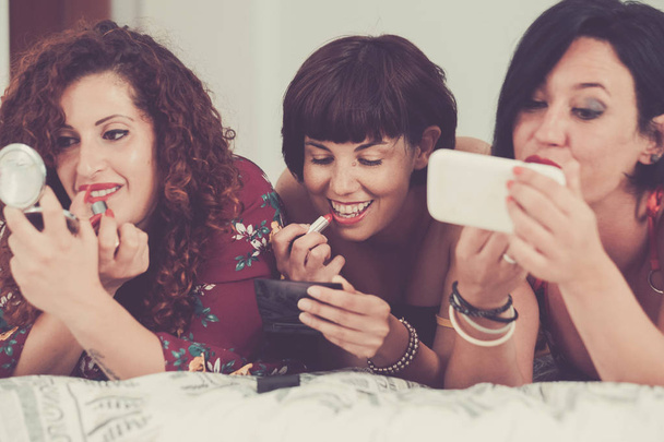 trzy kobiety pobyt przyjaciół razem położył się na łóżku, robi upewnij przed wyjść do noc stroną. pojęcie przyjaźni, jeden z nich korzystanie z telefonu komórkowego jak lustro aby sprawdzić jakość pracy - Zdjęcie, obraz