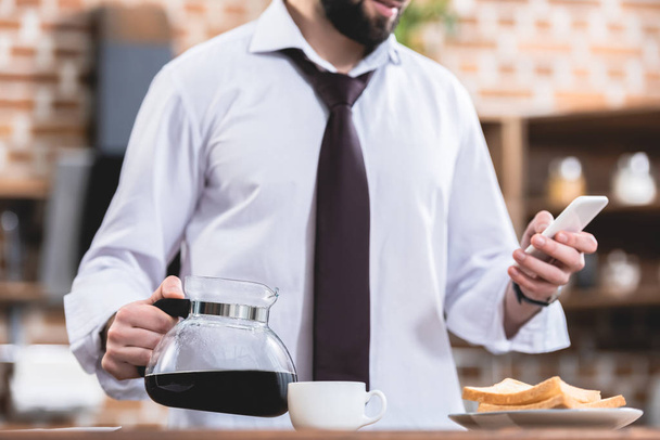 imagen recortada de hombre de negocios solitario sosteniendo la olla con café y teléfono inteligente en la cocina
 - Foto, imagen