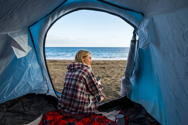 schöne blonde junge Frau tritt in das Zelt campen nur auf dem Sand des Strandes in der Nähe der Wellen des Ozeans. Nutzung ihres Mobiltelefons, um in freier Wildbahn verbunden zu bleiben - Foto, Bild