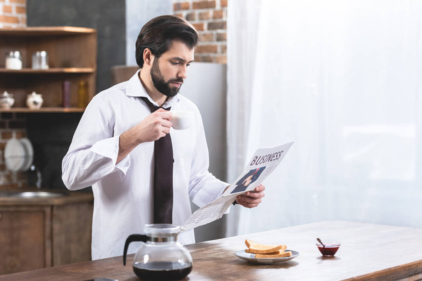 komea yksinäinen liikemies lukee sanomalehteä ja pitää kupin kahvia keittiössä
 - Valokuva, kuva