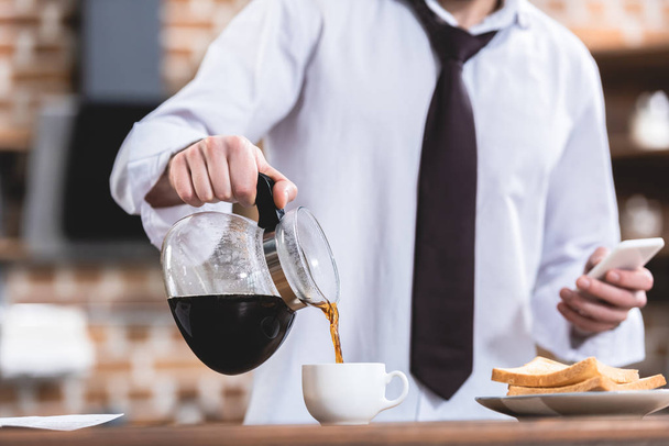 cropped image of loner businessman pouring coffee and holding smartphone at kitchen - Fotoğraf, Görsel