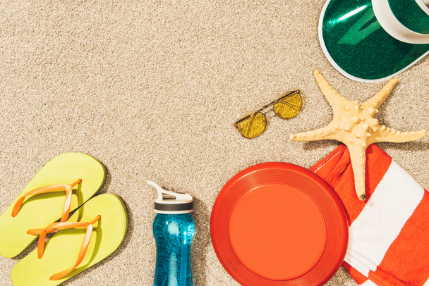 плоская лежала с фрисби, шапкой, флип-флопсом, солнцезащитными очками и бутылкой воды, расположенной на песке
 - Фото, изображение