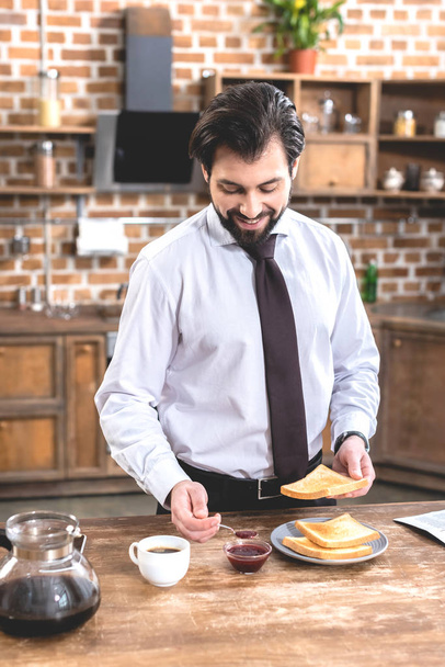 sonriente guapo solitario hombre de negocios añadir mermelada en tostadas en la cocina
 - Foto, Imagen