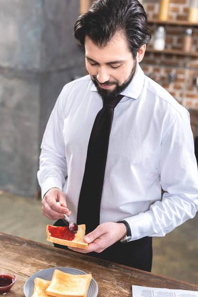 guapo solitario hombre de negocios añadiendo mermelada en tostadas en la cocina
 - Foto, Imagen