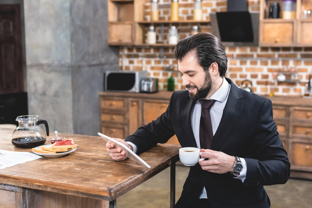 szép magányos üzletember látszó-on tabletta és a gazdaság csésze kávét, konyha - Fotó, kép