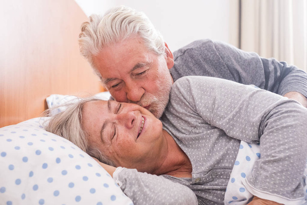 Couple of senior man and woman waking up and smiling with a hug while are in the bed at home. Vintage filter and light in the back. The man kiss the woman with love - Foto, Imagen
