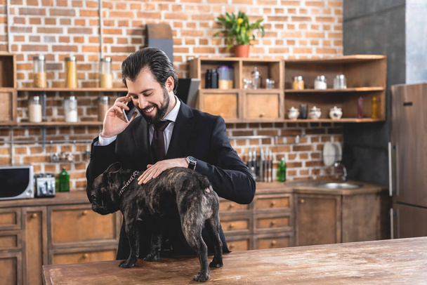 handsome loner businessman talking by smartphone and palming dog at kitchen - Foto, imagen