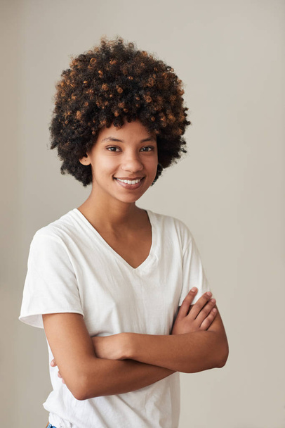 Retrato de una joven africana casualmente vestida con una afro sonriente mientras estaba de pie con los brazos cruzados en casa
 - Foto, imagen