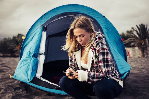 Always connected with technology blonde young woman stay ouside of her tent on the beach to enjoy freedom from work. - Photo, Image
