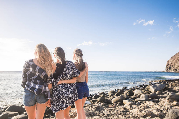 Group of three young nice girls embrace themselves looking at the ocean sunset in vacation. Team moment full of emotions and memories. Sunlight in a blue sky. - Photo, Image